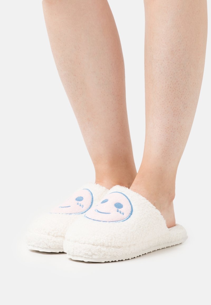 Jambes portant des chaussons blancs et duveteux avec une broderie d’un visage souriant sur le dessus sur un fond clair uni.