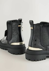Botines negros con acabado brillante, paneles laterales elásticos, acentos dorados y una suela gruesa. Patrón texturizado a lo largo de los lados resaltado.
