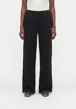 Personne portant un pantalon large noir avec taille élastique, un débardeur blanc et des chaussures foncées, debout devant un fond uni.