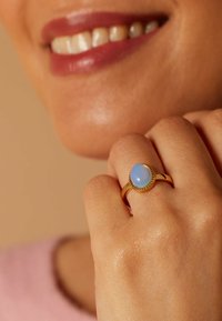 Gouden ring met een ovale blauwe opaal omgeven door een kralenaccent. De hand toont een gladde, gestructureerde huidskleur.