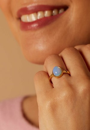 Gouden ring met een ovale blauwe opaal omgeven door een kralenaccent. De hand toont een gladde, gestructureerde huidskleur.