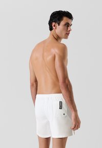 Fehér fürdőnadrág, amely könnyű anyagból k�észült, rugalmas derékkal, oldalzsebbel és fekete címkével a hátsó részén.