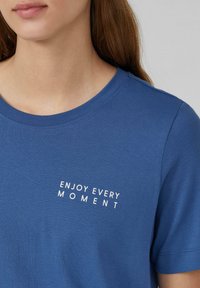 T-shirt en coton bleu avec un col rond et des manches courtes, portant un texte blanc "PROFITEZ DE CHAQUE MOMENT" positionné en diagonale sur le devant.