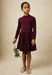 Bordo šaty s dlhými rukávmi, priliehavým bodom a nariadenou sukňou. Mäkká zamatová textúra. Zlaté baleríny s mašľou. Dieťa drží imelo.