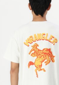 Biała bawełniana koszulka z dużym pomarańczowym i żółtym napisem "WRANGLER" oraz grafiką kowboja jeżdżącego na koniu z tyłu.