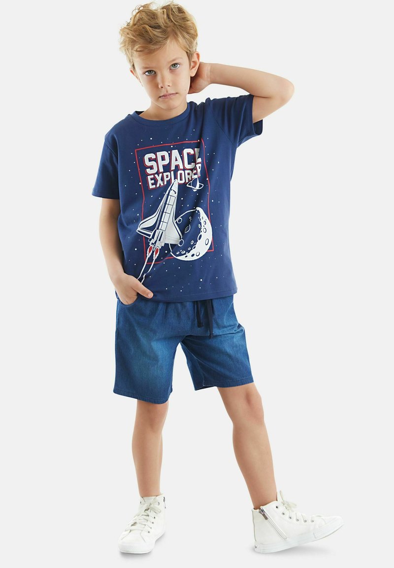Camiseta de algodón azul marino con gráfico de "EXPLORADOR DEL ESPACIO", combinada con pantalones cortos de mezclilla y zapatillas altas blancas.