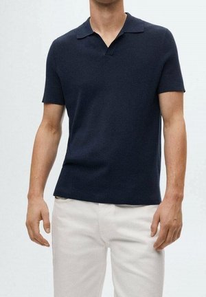 Homme portant un polo bleu marine à manches courtes associé à un pantalon blanc, debout devant un fond clair uni.