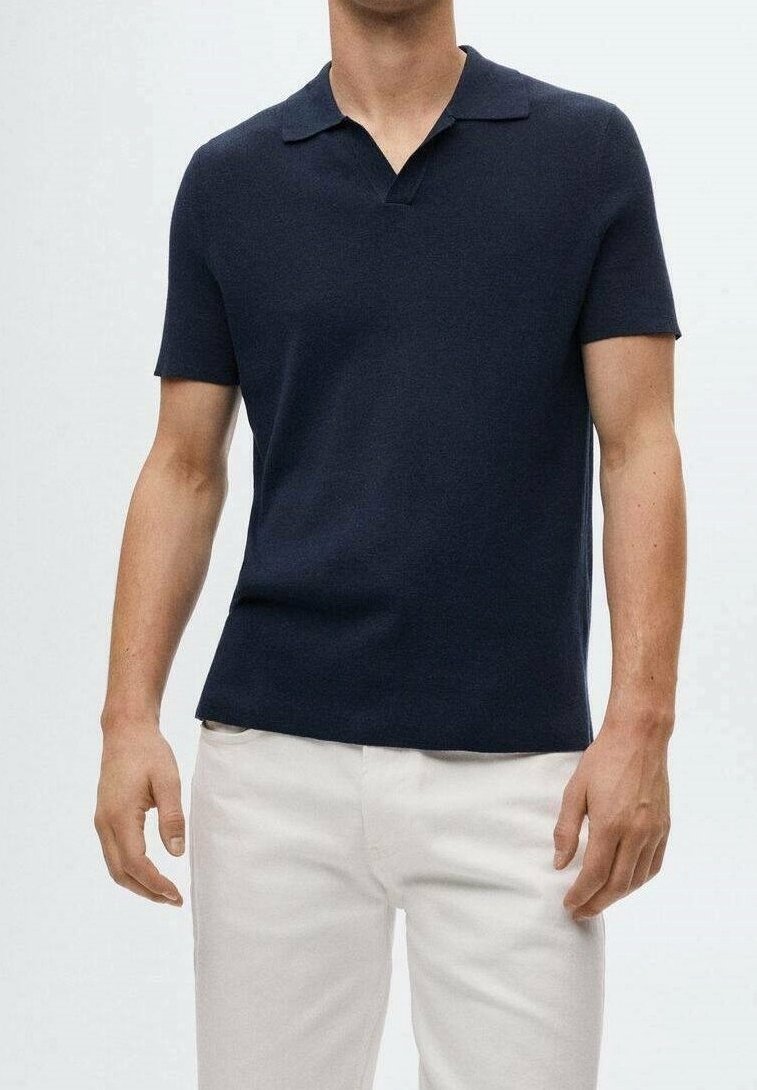 Homme portant un polo bleu marine à manches courtes associé à un pantalon blanc, debout devant un fond clair uni.