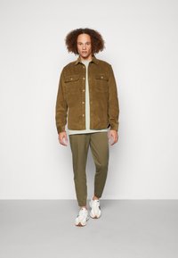 Chaqueta de camisa de pana marrón con dos bolsillos en el pecho, superpuesta sobre una camiseta verde clara, combinada con joggers oliva y zapatillas blancas.