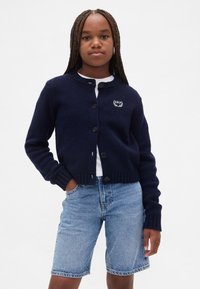 Modrý tmavě modrý pletený kardigan s kulatým výstřihem, sedmi knoflíky a vroubkovanými manžetami, s malým vyšitým logem na levé straně hrudi.