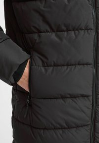 Melna puffa jaka ar izšūtu tekstūru, kurai ir rāvējslēdzēja kabata un elastīgas aproces, izgatavota no gludas, laika apstākļiem izturīgas auduma.