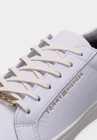 Gros plan d'une basket en cuir blanche avec des lacets beige et un logo Tommy Hilfiger doré sur le côté.
