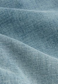 Lyseblåt denimstof med en skråvævning. Overfladen viser en glat tekstur med subtile variationer i farve og trådmønster.