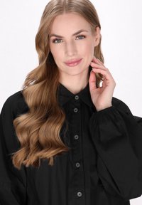 Černá košile na knoflíky s límečkem, dlouhými volnými rukávy a hladkým povrchem. Model má vlnité vlasy, které splývají dolů.