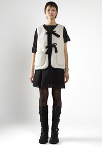 Weißer Fleece-Weste mit schwarzem Rand und Bandbändeln, getragen über einem schwarzen Kleid mit durchsichtigen Ärmel. Das Modell trägt kniehohe schwarze Stiefel mit Schnallen.