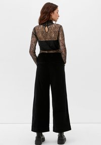 Schwarzes Spitzenoberteil mit hohem Kragen und Rückenschlitzdetail, kombiniert mit weiten schwarzen Hosen. Glatte Textur und lockere Passform über das gesamte Outfit.