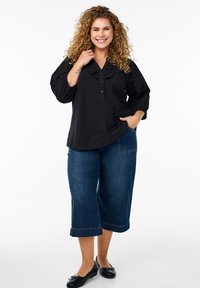 Schwarze Bluse aus Stoff mit Rüschenausschnitt, Knopfdetails und drei Viertelärmeln, kombiniert mit blauen Jeans-Culottes und schwarzen flachen Schuhen.