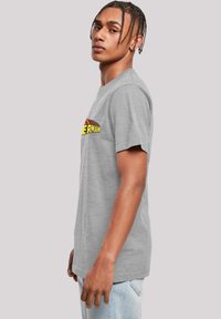 Šedé bavlněné tričko s žlutou a červenou grafikou "SUPER MAN" na přední straně, krátkými rukávy a uvolněným střihem.