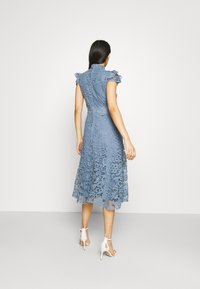 Niebieska koronkowa sukienka z krótkimi falbaniastymi rękawami, dopasowaną górą i zwiewną spódnicą. Szczegółowy wzór koronkowy, długość do kolan, noszona z białymi obcasami.