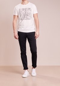 T-shirt blanc à manches courtes avec imprimé graphique noir, associé à un pantalon slim noir et des baskets blanches, debout sur un sol clair.