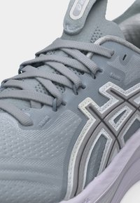 Κοντινή λήψη ενός γκρι αθλητικού παπουτσιού με υφασμάτινη πλέξη, σταυρωτά κορδόνια και λευκό-γκρι λογότυπο με σχέδιο στην πλευρά.