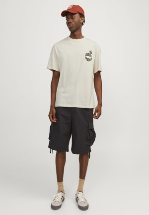 Ανοιχτό μπεζ βαμβακερό μπλουζάκι με γραφικό "ΘΕΩΡΙΑ ΧΑΟΣ" στο αριστερό στήθος, συνδυασμένο με μαύρες cargo βερμούδες, μπεζ κάλτσες και γκρι αθλητικά παπούτσια.