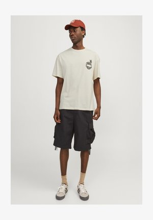 Kerge beež puuvillane T-särk „KAOS TEORIA” vasakus rinnas, kombineeritud mustade cargo lühikesed püksid, beežide sokkide ja hallide spordijalatsitega.