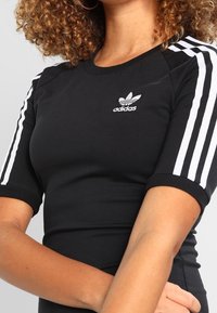 Černé bavlněné tričko s krátkým rukávem, na každém rukávu se třemi bílými pruhy a malým bílým logem Adidas na hrudi.