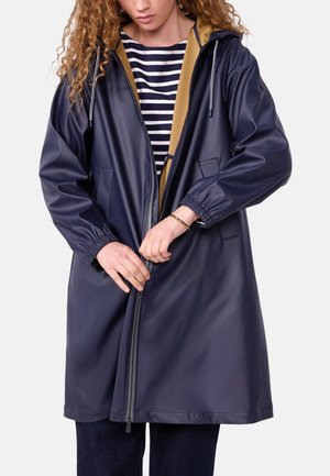 Person, die einen dunkelblauen Regenmantel über einem gestreiften Shirt zuzieht, mit den Händen am Reißverschluss und teilweise sichtbarem hellbraunem Haar.