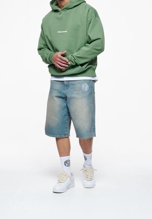 Sudadera con capucha verde con logotipo en el frente, pantalones cortos de mezclilla azul claro, calcetines blancos y zapatillas blancas con cordones gruesos. Estilo informal de ropa urbana.