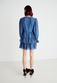 Vestido mini em camadas azul, feito de tecido texturizado, com mangas bufantes e fecho de correr nas costas. Combinado com sapatos de salto alto adornados com grandes laços pretos.
