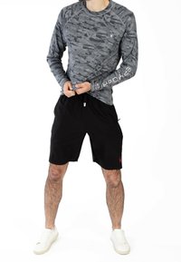 Camicia a maniche lunghe grigia in mimetica con tessuto testurizzato e un logo sulla manica, abbinata a pantaloni corti neri con tasche laterali e cordoncino.