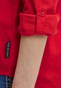 Rotes Hemd mit Knöpfen, hochgerollten Ärmeln und einem einzigen Knopfverschluss, versehen mit dem Etikett "DOWN THE RIVER" nahe dem Saum. Strukturiertes Gewebe.