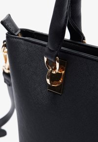 Sac à main noir texturé avec des anneaux en métal doré attachant les poignées, doté d'une fermeture à languette et d'une bandoulière sur un fond blanc.