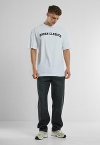 Balta medvilnė marškinėliai su juodu užrašu "URBAN CLASSICS", derinami su tamsiai pilkomis džinsais ir šviesiai rudos sportinėmis bateliais su juodais akcentais.