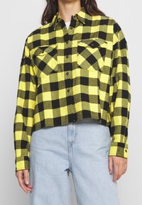 Woman wearing yellow and black checkered button-up shirt with front pockets, paired with light blue high-waisted jeans.