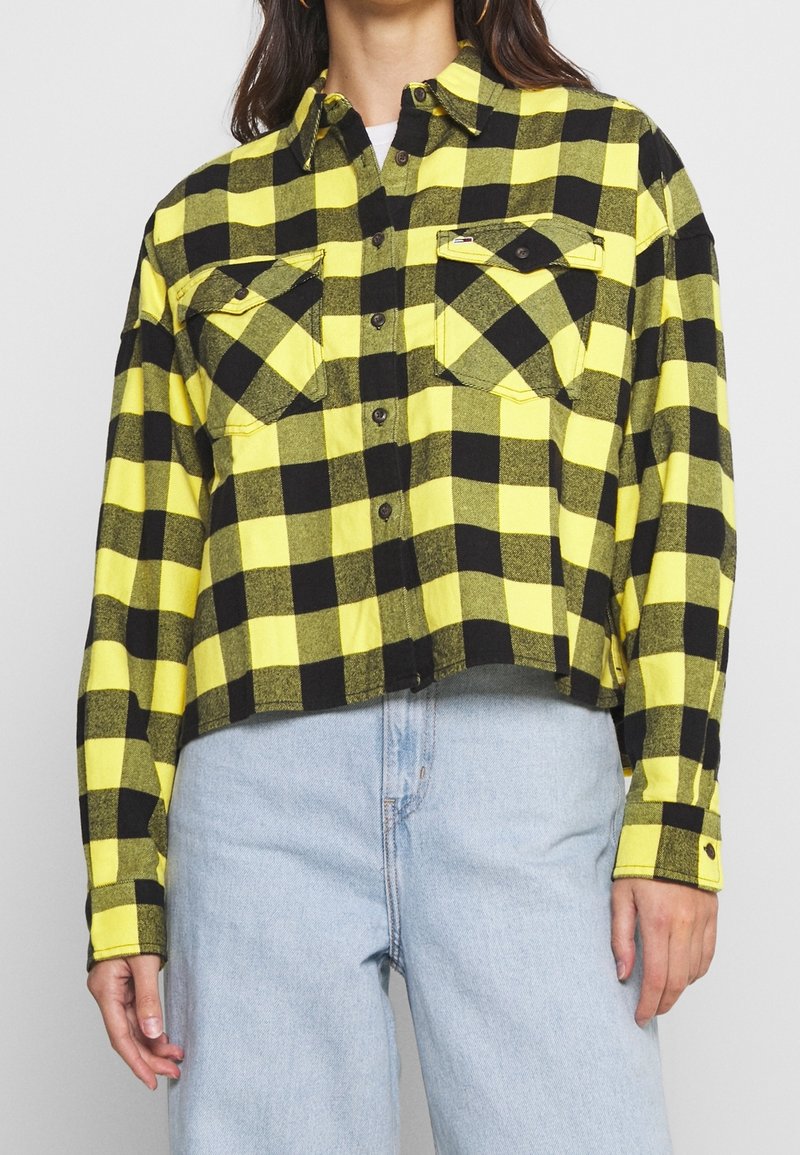 Woman wearing yellow and black checkered button-up shirt with front pockets, paired with light blue high-waisted jeans.