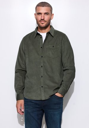 Mann mit kurzen Haaren und Bart trägt ein dunkelgrünes langärmliges Hemd über einem weißen T-Shirt und dunkelblauen Jeans, steht mit einer Hand in der Tasche.