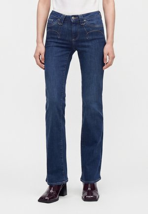 Personne portant un jean droit bleu foncé et des bottines bordeaux foncé à bout carré sur fond blanc.