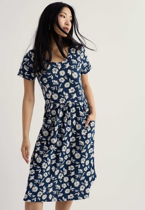 Femeie cu păr negru și lung, purtând o rochie bleu închis până la genunchi, cu modele florale albe, stând cu o mână în buzunar.