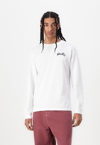 White long-sleeve shirt made of cotton with a round neckline and black logo on the left chest. Model wearing light burgundy pants.