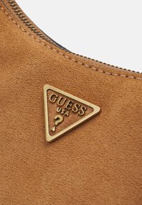 Primer plano de un material de gamuza marrón claro con el logo triangular dorado de Guess USA y costuras visibles cerca de una cremallera.