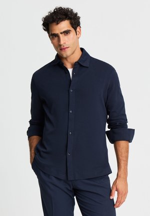 Uomo con capelli ricci che indossa una camicia blu scuro abbottonata con le maniche arrotolate e pantaloni blu scuro abbinati, in piedi davanti a uno sfondo uniforme.