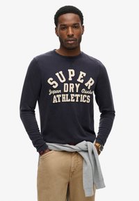 Marineblå langærmet t-shirt med cremefarvet tekst, der står "SUPER DRY ATHLETICS", og strikket tekstur; stylet med en grå sweater bundet om livet.