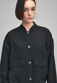 Zwarte button-up jas met een mandarin kraag, gouden knopen en twee borstzakken; gemaakt van glad, gestructureerd weefsel.