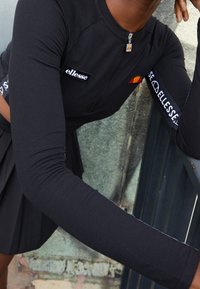 Svart långärmad topp med dragkedja vid kragen, med logotyper på bröstet och ärmen. Slät tyg och kroppsnära design.