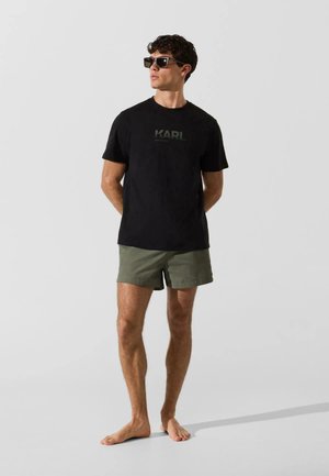 Férfi fekete "KARL" feliratú pólóban, olívazöld rövidnadrágban és napszemüvegben, mezítláb áll egy sima világos háttér előtt.