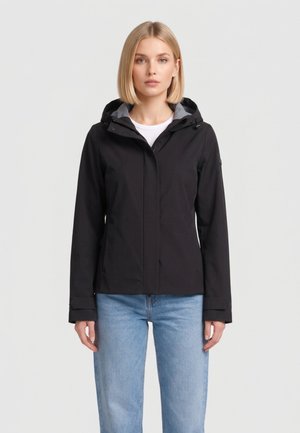 Donna bionda che indossa una giacca nera con cappuccio e jeans azzurri chiari, in piedi davanti a uno sfondo uniforme grigio chiaro.