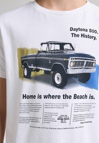Hvid bomulds T-shirt med grafik af en vintage sort pickup-truck med blå og grønne accenter, ledsaget af tekst om Daytona 500.