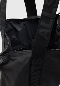 Black tote bag with smooth fabric, zip closure, and large interior. Features bold logo straps and small pocket on the side. Minimalist design.